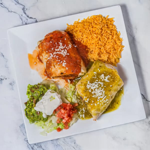 Two burritos, one with red sauce and one with green, served with rice, guacamole, and sour cream.